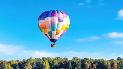 Obraz premium A colorful hot air balloon floating gracefully in the sky above a lush green forest, vibrant autumn foliage, clear blue sky, and serene adventure concept.