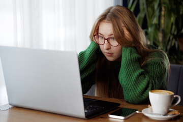Stressed woman with glasses looking at laptop in cafe, showing signs of frustration.