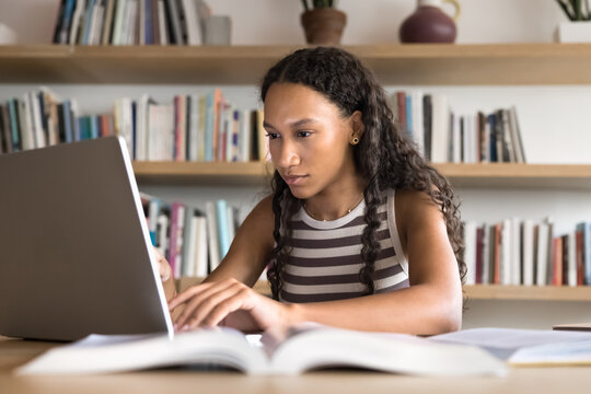 Blended education. Busy female student focused on self training at library do academic research work surrounded by books type on notebook succeeding in combining offline studying with digital learning