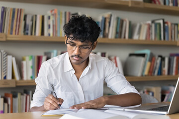 Engaged in learning. Serious young male student in glasses do academic assignment during self study time in library work to earn higher education take notes by hand from printed books notebook screen