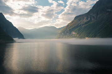 View of the entrance to the Geirangerfjord at sunrise with fog on the surface and mountains in the background in Norway.