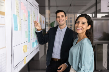 Two happy male and female business colleagues standing together at scrum board with sticky notes,...