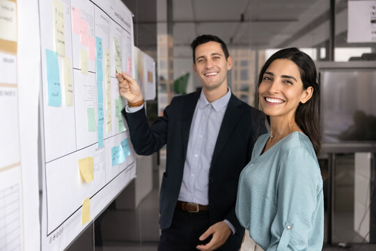 Two happy male and female business colleagues standing together at scrum board with sticky notes, looking at camera, smiling, posing for corporate portrait, enjoying teamwork, collaboration - Powered by Adobe