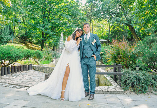 A lovely wedding couple in a scenic outdoor location. The bride, wearing a beautiful white dress, lays her head on the groom&rsquo;s shoulder, while the groom is in an elegant suit.