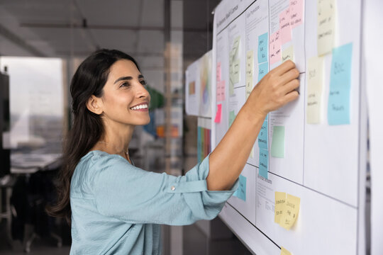Successful young Latin project manager woman working on tasking with sticky notes on board, using scrum management method, planning business process and moving stickers, smiling