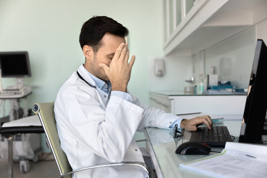 Doctor sit at desk with computer and papers, cover face with hand, feel exhausted, overloaded, looks stressed due to medical errors or misdiagnosis, make mistake, dealing with difficult patient case