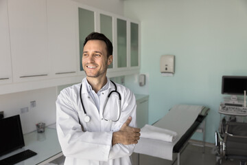 Happy Latin man doctor in white coat standing confidently in contemporary consulting room, looking...