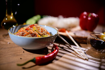 Colorful bowl of noodles with fresh vegetables and spices on wooden table during evening setting