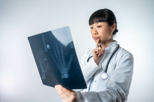 Beautiful thoughtful Asian female doctor looking at hand X-ray image. Medical professional analyzing radiograph isolated on white background.
