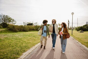 Fototapeta premium Group of three friends walking and talking while laughing and holding a bag, package, and ball, outdoors, on a walk