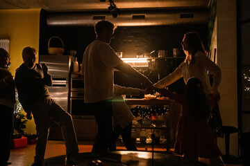 Father, mother, daughter, and son are dancing round dances while grandfather and grandmother dance next to them, in the kitchen, at Christmas
