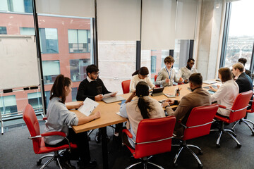 A group of nine employees and a male intern are sitting at a table in a meeting, in an office, during an internship