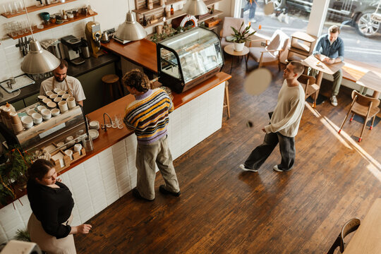 A male employee stands behind the bar while two customers and a female owner walk across from him, in a cafe