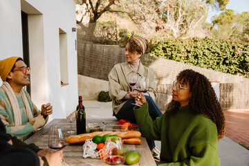 A group of three friends are sitting at a table and talking while a female friend sits next to them at the table, they are holding glasses, outdoors, during dinner