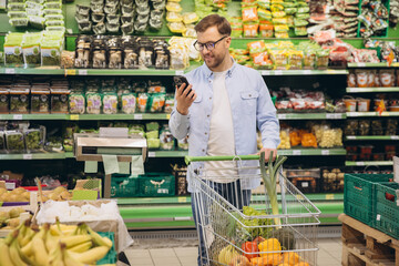 Smiling customer using phone and pushing shopping cart in supermarket