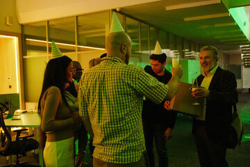 A male employee stands and smiles as he prepares to clink glasses with a male employee standing next to him while a group of six employees stand next to them, in the hall, at a retirement party