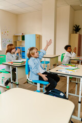 Obraz premium A group of three schoolchildren sit at desks and look ahead while one of them raises his hand, at school, in the classroom