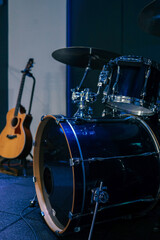 Close-up of a dark blue drum set on a stage, with a wooden acoustic guitar in the background, under...