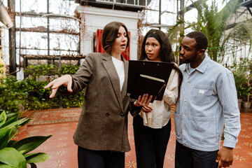 A man and a woman are standing and looking at a woman organizer who is standing next to them and talking while looking at a clipboard she is holding, in a venue, preparing for a wedding