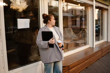 Female owner standing and holding a cup and computer tablet while looking away, on the street,...