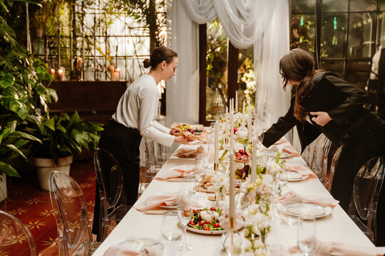 A female organizer stands and holds a clipboard while holding her hand near the table on which a female waiter places a plate, standing opposite her, preparing for a wedding party