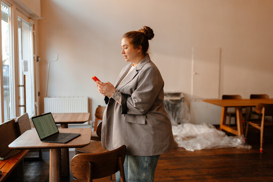 The female owner is standing and smiling while typing on the phone she is holding in front of her, preparing for the opening of the cafe - Powered by Adobe