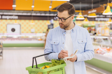 Focused man writing grocery list in supermarket with shopping basket