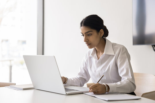 Serious focused Indian woman using laptop, making notes, writing down important information, studying or working on-line using internet and educational business platform. Workflow, modern tech usage