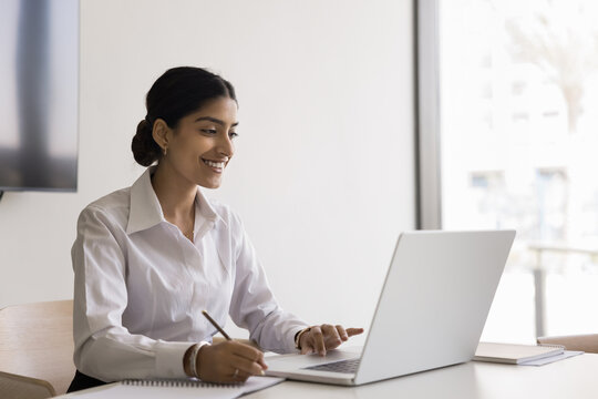 Attractive smiling Indian woman sitting at table with laptop, holds pen, jotting information, making important notes, focused in learning or working process using new program, app, website or platform