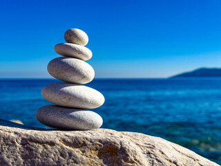 Fototapeta premium CLOSE UP: Stack of five smooth white pebbles is perfectly balanced on a stone surface by the sea. Calm blue ocean wavy and clear sky create a tranquil summer backdrop, evoking peace and mindfulness.