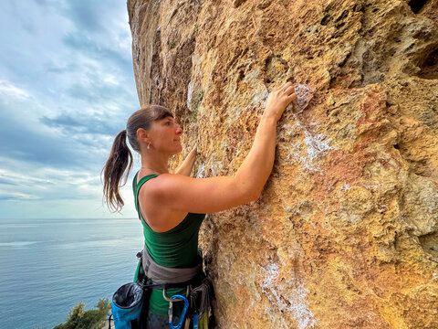 CLOSE UP, PORTRAIT: Woman climbs a vertical limestone cliff above the Aegean Sea in Greece. Focused female climber is dealing with a challenging climbing route and searches for her next handhold.