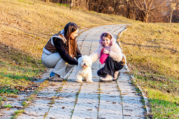 A picture in the lap of nature on a spring background with lots of very colorful and beautiful colors, two girls were playing with their best friend, the little white dog, who is the happiest to have