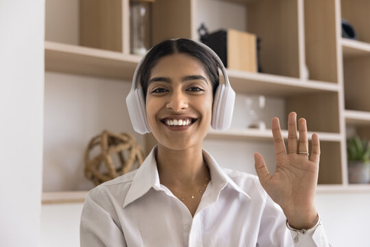 Head shot of young Indian woman wear wireless headphones engaged in virtual meeting, take part in business-related or personal conversation using video call application. Tutor greets learner, tuition