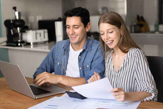 Happy wife and husband sitting at table in modern kitchen, sorting out financial papers, paying utility bills, smiling, feel satisfied with incomes and earnings, enough finances for monthly payments