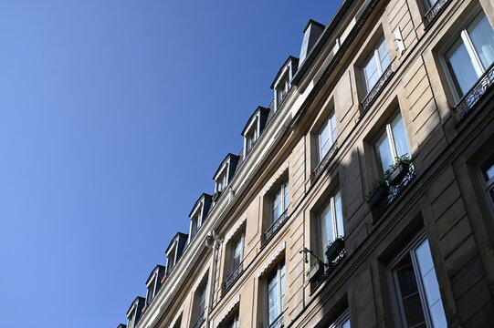 Elegant Parisian residential building captured from a low angle against a clear blue sky. Classic Haussmann-style architecture with tall windows, wrought-iron balconies, and beige stone fa&ccedil;ade. Bright
