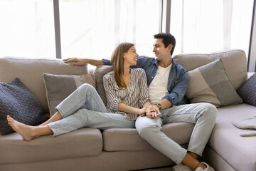 Obraz premium Relaxed couple sitting comfortable on couch, smiling and lead talk spend pastime together in contemporary living room. Young homeowner family resting on cozy sofa, enjoy conversation on weekend indoor