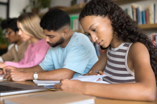 Self training. Busy teen female high school student engaged in studying at library with group of diverse multicultural peers use paper workbooks notebook to search information prepare for lesson exam