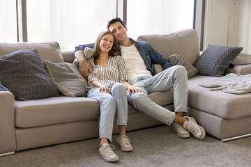 Obraz premium Beautiful Latin couple sitting on comfortable couch in modern living room, looking at camera with smile, enjoy relaxation at rented apartment. Homeowners, newlyweds home portrait. Bank loan, new house