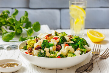 Steamed Broccoli Cauliflower Salad with kalamata olives, red bell pepper, toasted almond slices in white bowl with brick wall, fresh basil and lemon in background, horizontal view from above, close-up
