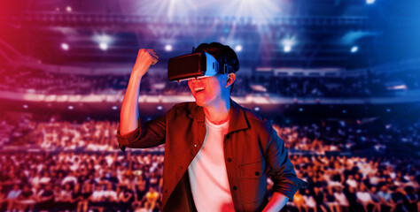 Asian man wearing VR glasses and casual cloth and surrounded with crowds cheering and sitting at arena. Gamer celebrating winning in video game while using augmented reality technology. Ingenuity.