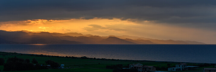 Golden rays break through dark clouds over tranquil sea and distant mountains at dusk