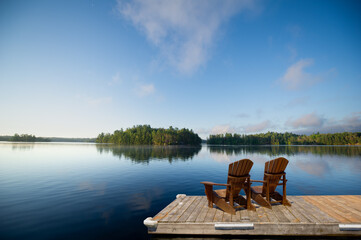 Two Adirondack chairs sit on a wooden dock overlooking a calm Muskoka lake, with fluffy clouds and...