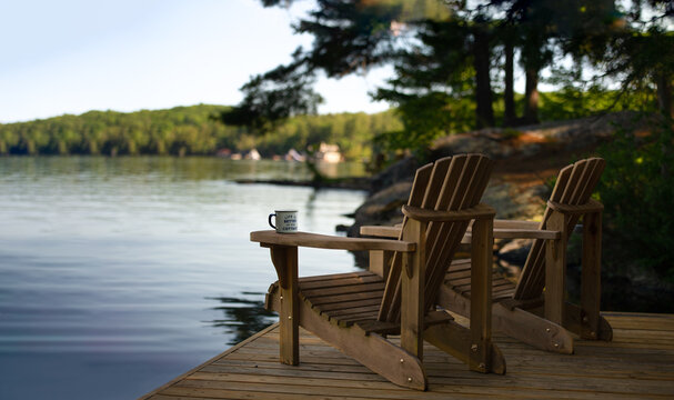 Two Adirondack chairs sit on a cottage dock, facing a calm Muskoka lake with cabins in view on a sunny summer morning. A coffee tin on one arm reads "Life is better at the Cottage".
