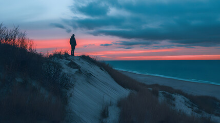 Obraz premium Silhouette on the Dunes: A solitary figure stands atop a sandy dune, silhouetted against a vibrant sunset over the ocean. Peaceful contemplation at twilight.