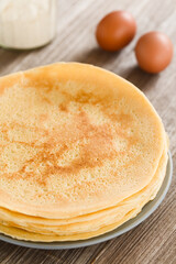 Stack of fresh homemade crepes, thin French-style pancakes, on plate with flour and eggs in the back (Selective Focus, Focus one third into the image)