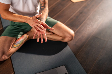 A young yoga instructor is conducting an online class while sitting on a yoga mat in his studio. He explains to his student using hand gestures.