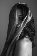 Naklejka premium A grayscale portrait captures a woman with a draped fabric over her head, partially obscuring her face. Her gaze is direct, creating a sense of mystery. Soft light emphasizes her features.