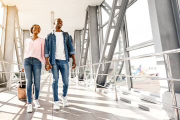 Fototapeta premium Travelling Together. Cheerful Black Couple Holding Hands While Walking In Airport Terminal, Happy African American Spouses Carrying Luggage Suitcases While Going To Boarding Gate, Copy Space