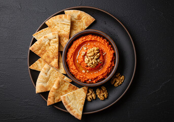 Top view of roasted red pepper and walnut dip in a rustic stoneware bowl, surrounded by crispy toasted pita chips. Presented on black background in high-res, professionally styled.