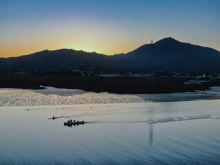 A motorboat carries passengers on a river in Larkspur, California, USA at sunset. A lone rower is...